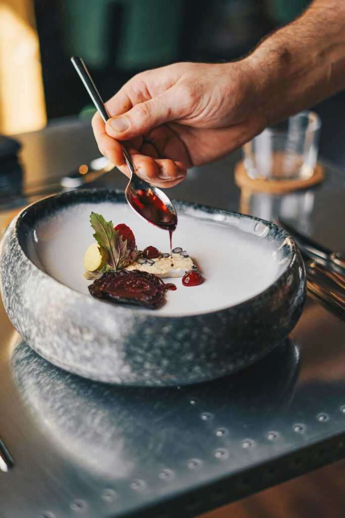 A close-up of a hand carefully drizzling sauce over a neatly plated dish in a stone bowl, with minimal portions arranged thoughtfully and a soft, blurred dining setup in the background, highlighting the precision and artistry of food presentation.