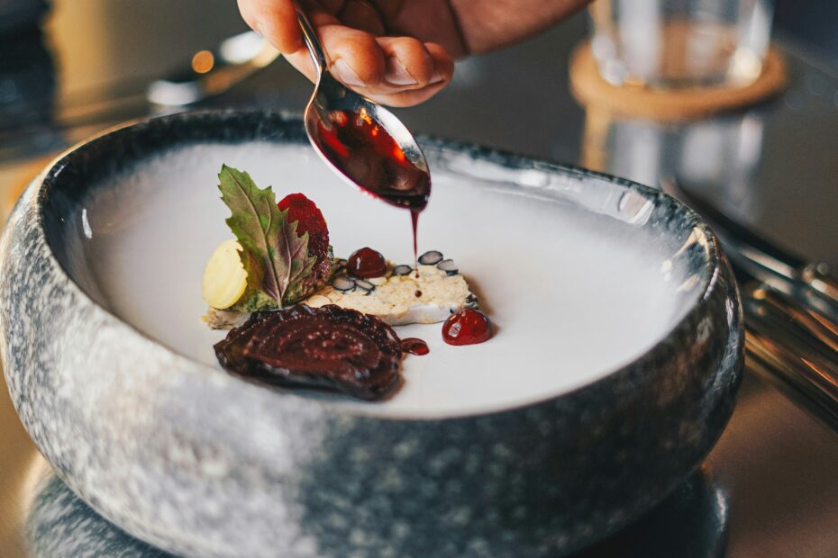 A close-up of a hand carefully drizzling sauce over a neatly plated dish in a stone bowl, with minimal portions arranged thoughtfully and a soft, blurred dining setup in the background, highlighting the precision and artistry of food presentation.