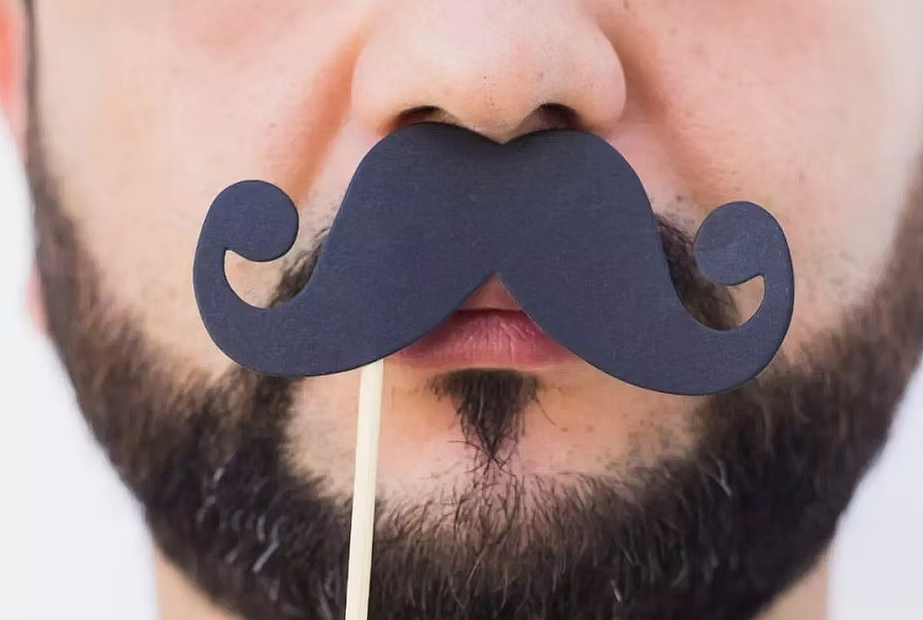Close-up of a man’s face holding a curled, paper moustache prop on a stick over his lips, with a trimmed beard visible—playful representation of Movember moustache culture.