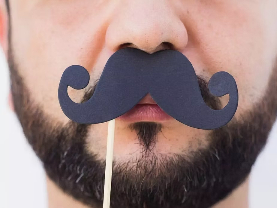 Close-up of a man’s face holding a curled, paper moustache prop on a stick over his lips, with a trimmed beard visible—playful representation of Movember moustache culture.