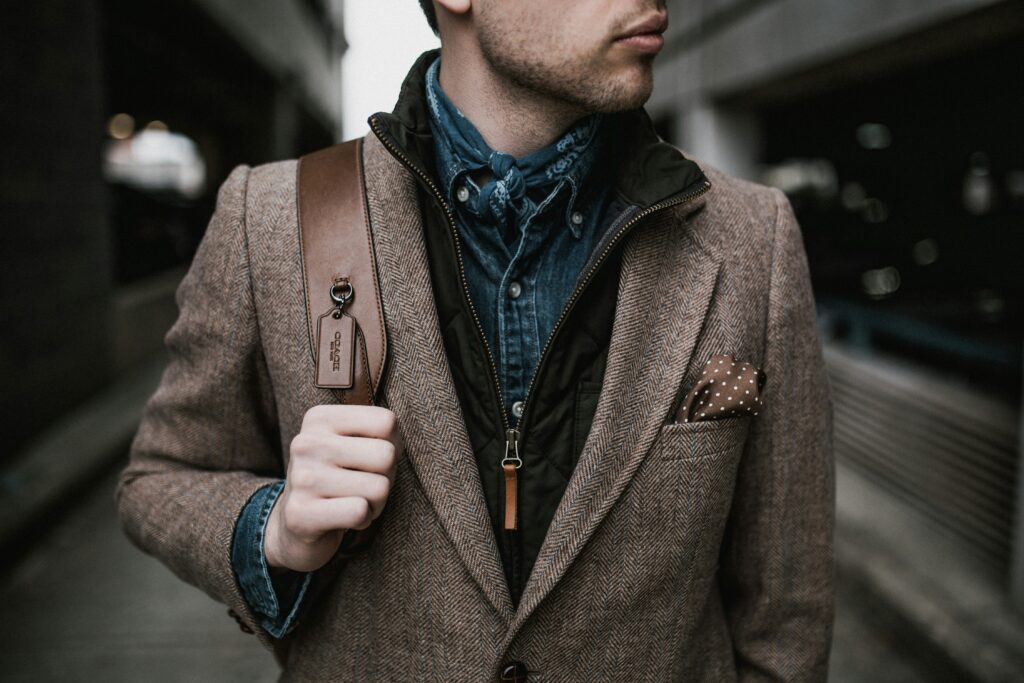 A man wearing a brown herringbone blazer layered over a denim shirt and black quilted vest, styled with a neckerchief, pocket square, and leather shoulder bag, standing in an urban setting.