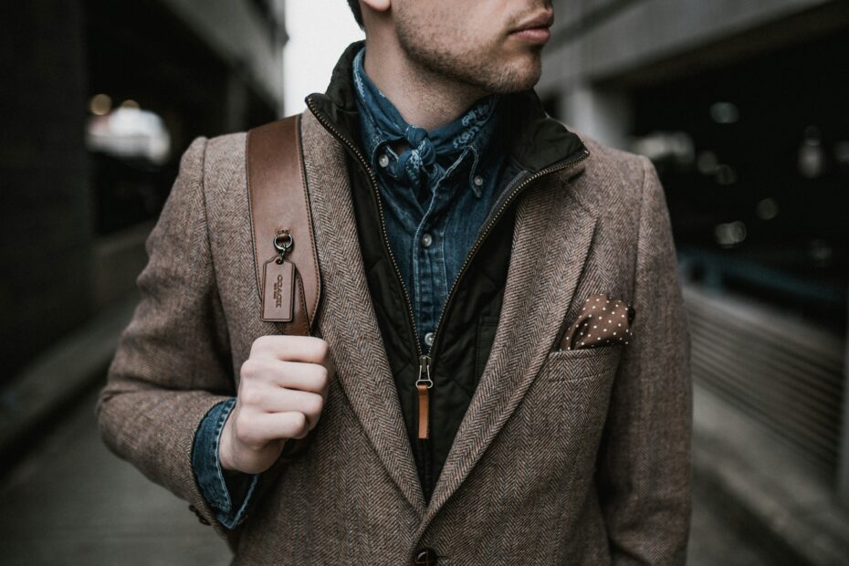 Man wearing a brown herringbone blazer layered over a denim shirt and black quilted vest, styled with a neckerchief, pocket square, and leather shoulder bag, standing in an urban setting.