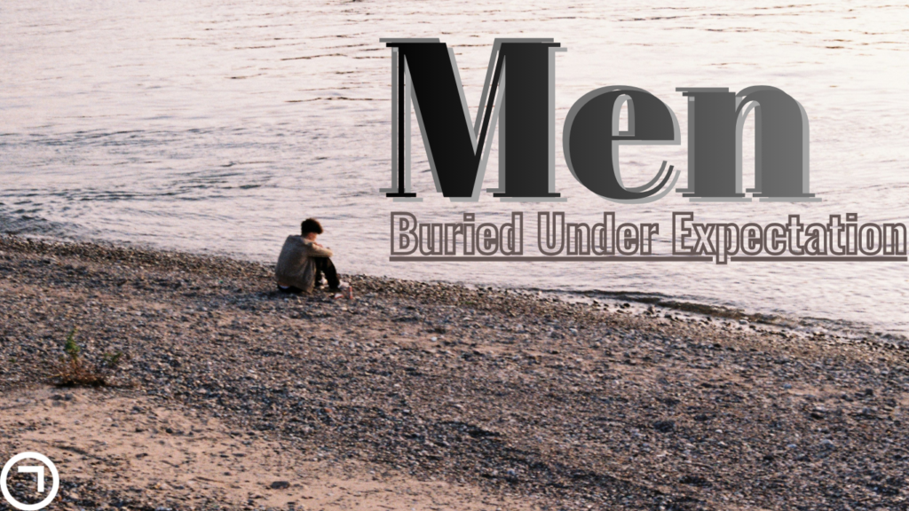 A lone man sits on a quiet shoreline, hunched and isolated, with the words “Men: Buried Under Expectations” overlaid—capturing the weight of societal pressure and emotional solitude.