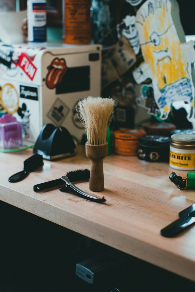 Wooden beard brush with light bristles placed on a grooming table, surrounded by barber tools and styling products in a casual barbershop setting.