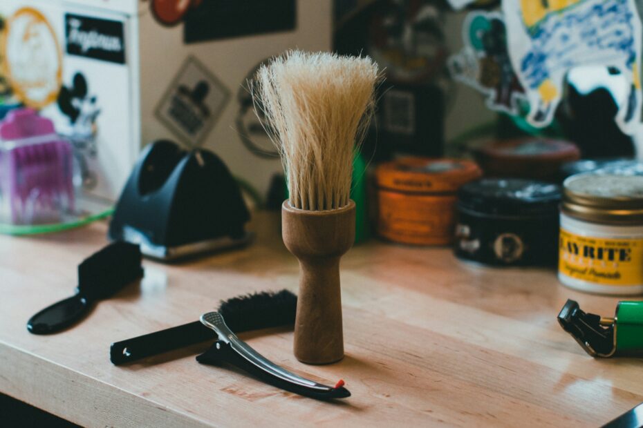 Wooden beard brush with light bristles placed on a grooming table, surrounded by barber tools and styling products in a casual barbershop setting.