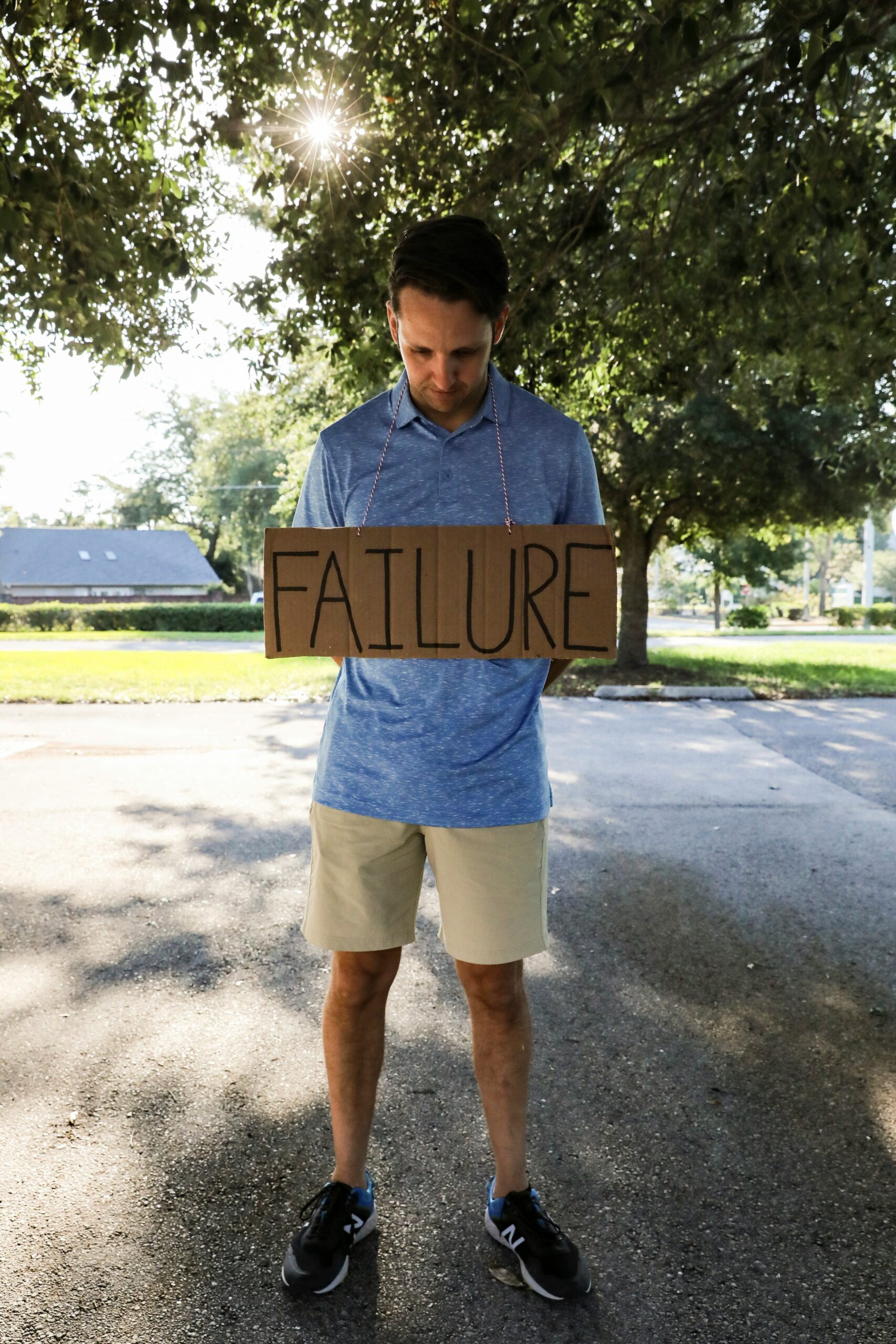 A man standing outdoors under a tree, looking down with a cardboard sign hanging around his neck that reads “FAILURE.”