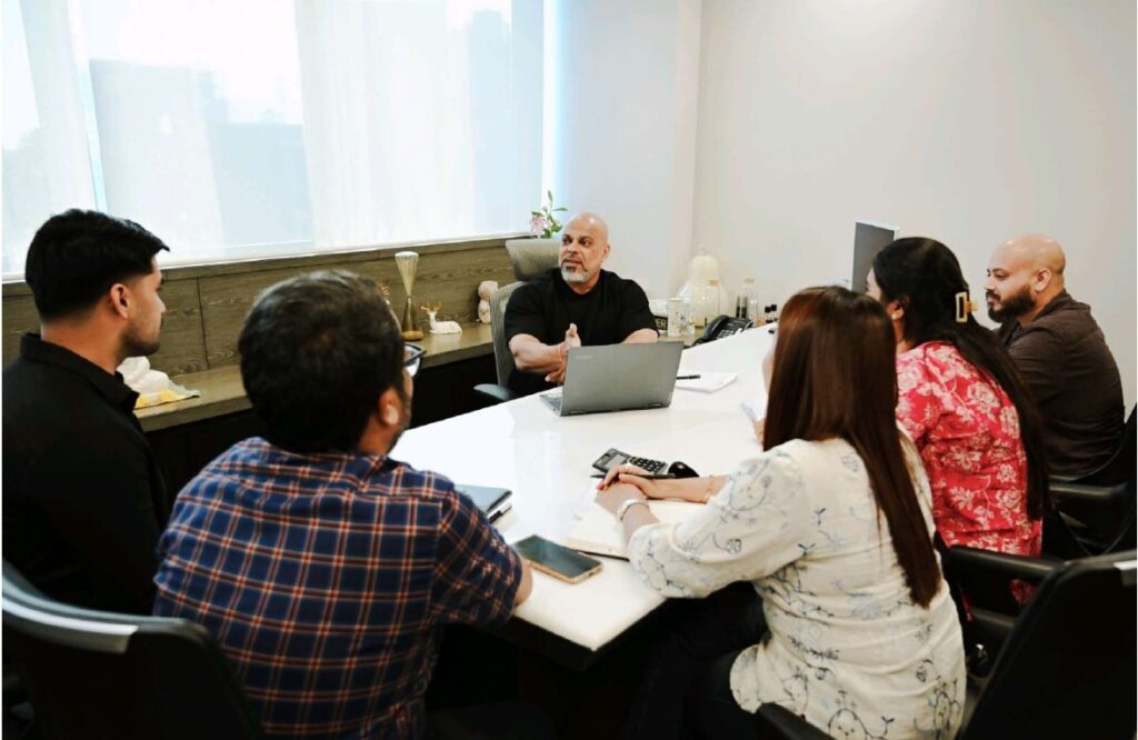Business team meeting in a modern office conference room, with a leader presenting ideas to colleagues seated around a table with laptops and notebooks.