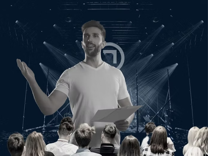 A charismatic man in a white t-shirt stands on a stage, gesturing with an open hand while addressing an attentive audience. The background features dramatic stage lighting and a dark, modern setting, symbolizing effective public speaking and storytelling.