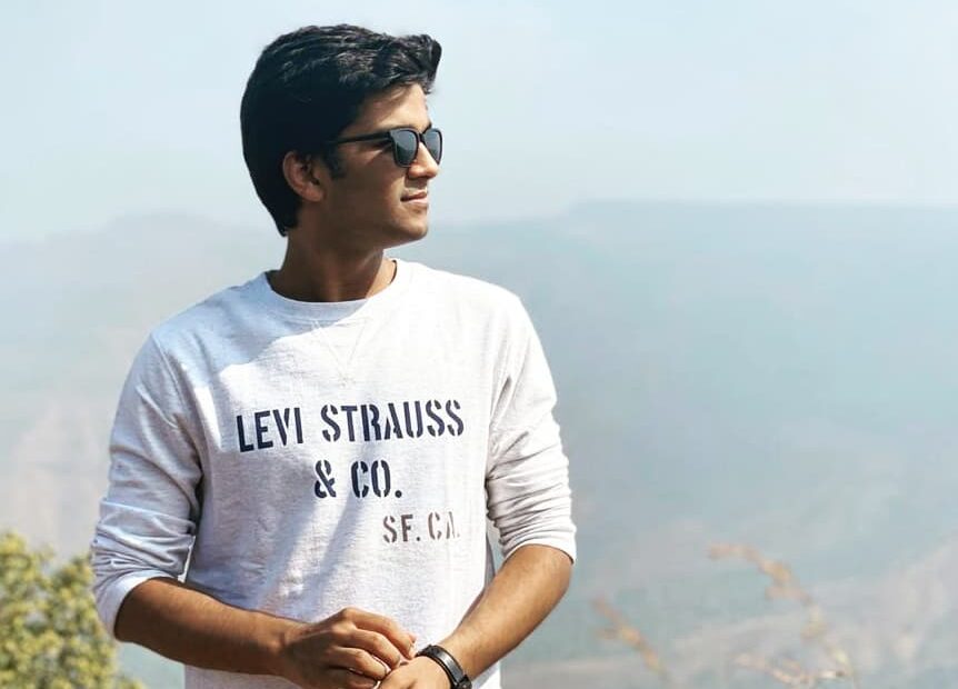 Young man wearing sunglasses and a white Levi Strauss & Co. sweatshirt, standing outdoors against a scenic mountain backdrop on a sunny day.