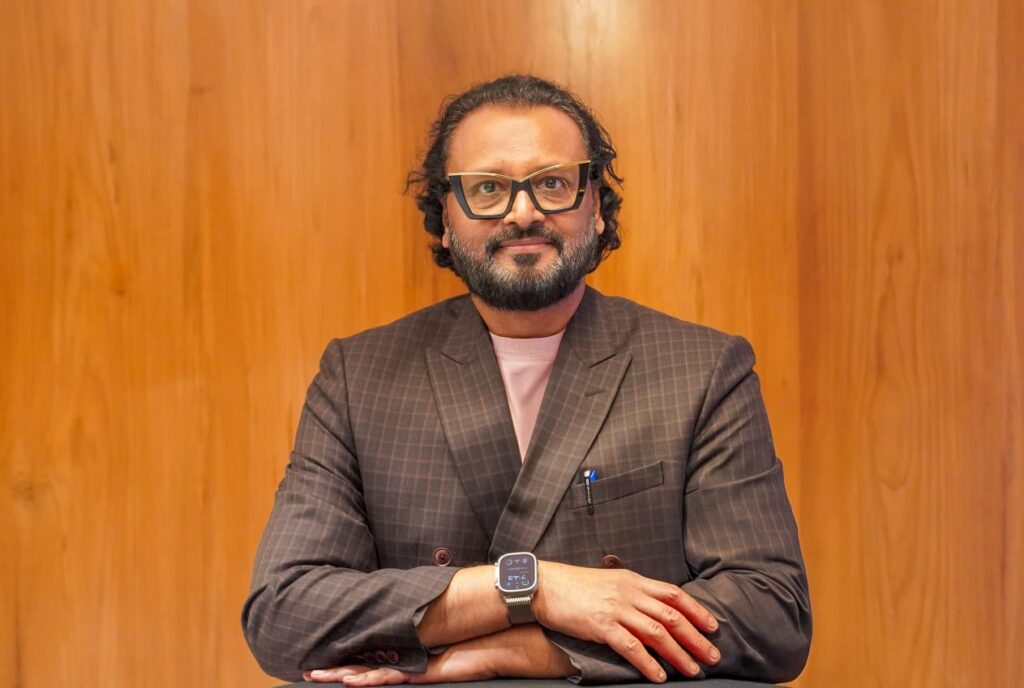 a bearded man with wide forehead smiling wearing spectacles and a smart watch with brown background