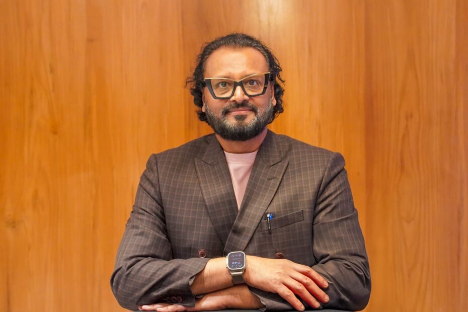 a bearded man with wide forehead smiling wearing spectacles and a smart watch with brown background