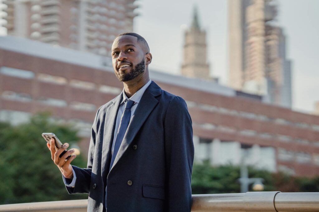 A smiling young male professional in suit holding a smartphone with the right hand, looking at the sky.