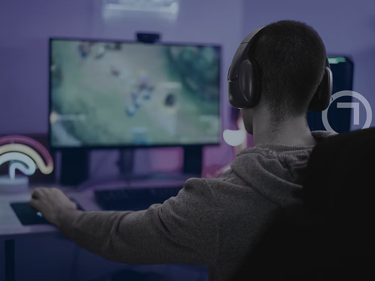 Person wearing headphones while gaming on a desktop computer in a dimly lit room with a blurred game screen in the background.