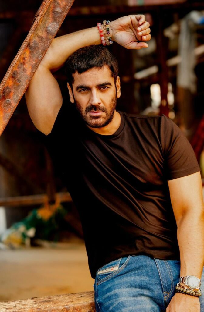Arjan Bajwa leans casually against a metal structure in an industrial setting, dressed in a fitted black T-shirt and blue jeans. His intense gaze and relaxed pose create a powerful, contemporary portrait highlighting his charismatic screen presence.