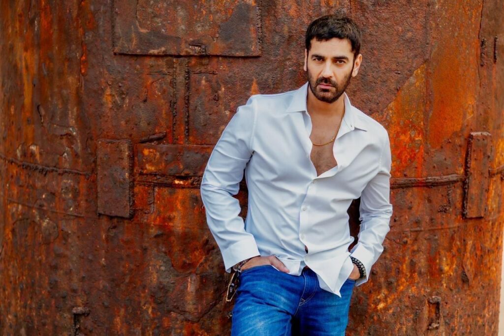 Arjan Bajwa poses against a weathered rust-colored industrial backdrop, wearing a crisp white shirt and blue jeans. With a confident expression and relaxed stance, the actor brings a rugged yet sophisticated charm to the portrait.
