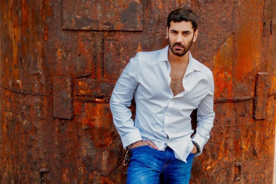 Arjan Bajwa poses against a weathered rust-colored industrial backdrop, wearing a crisp white shirt and blue jeans. With a confident expression and relaxed stance, the actor brings a rugged yet sophisticated charm to the portrait.