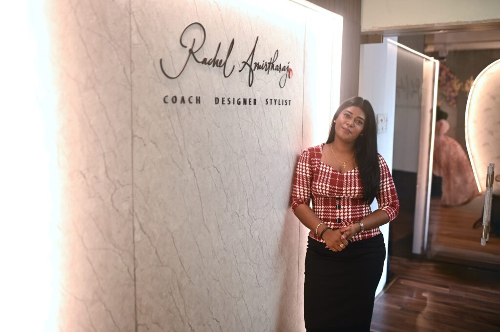 Rachel J Amirtharaj, founder and fashion designer of La Fantaisie, standing inside her design studio beside a branded wall showcasing her role as coach, designer, and stylist.
